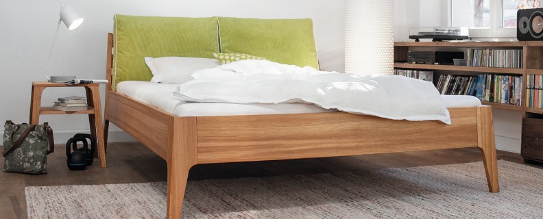 Bettgestell Kouro der Firma Sira. Bett mit einem Holzrahmen und Polsterkopfteil. Kissen in grün.